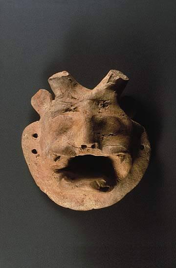 In the center is miniature mask of horned deity with human face and bared teeth of a tiger. A large mustache or divided upper lip frames the canines, and a flaring beard adds to the effect of rage. The eyes are defined as raised lumps that may have originally been painted. Short feline ears contrast with two short horns similar to a bull rather than the curving water buffalo horns. Two holes on either side allow the mask to be attached to a puppet or worn as an amulet.
