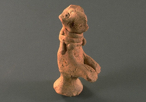 Figurine of begging dog with upraised front paws and wearing a beaded collar. The back legs have been shaped into a stand. Hand formed with applique ornaments and eyes.