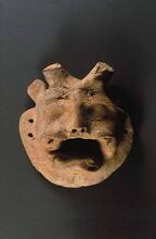 In the center is miniature mask of horned deity with human face and bared teeth of a tiger. A large mustache or divided upper lip frames the canines, and a flaring beard adds to the effect of rage. The eyes are defined as raised lumps that may have originally been painted. Short feline ears contrast with two short horns similar to a bull rather than the curving water buffalo horns. Two holes on either side allow the mask to be attached to a puppet or worn as an amulet.