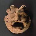 In the center is miniature mask of horned deity with human face and bared teeth of a tiger. A large mustache or divided upper lip frames the canines, and a flaring beard adds to the effect of rage. The eyes are defined as raised lumps that may have originally been painted. Short feline ears contrast with two short horns similar to a bull rather than the curving water buffalo horns. Two holes on either side allow the mask to be attached to a puppet or worn as an amulet.